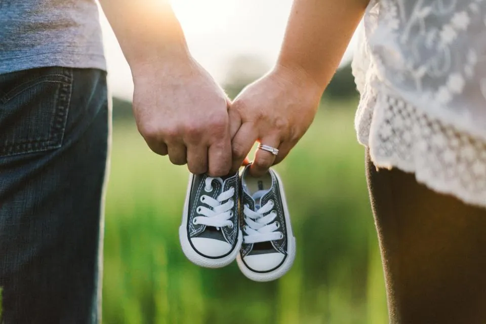 Un couple, cadré à hauteur de mains, tient ensemble une paire de petites baskets de bébé dans un pré.