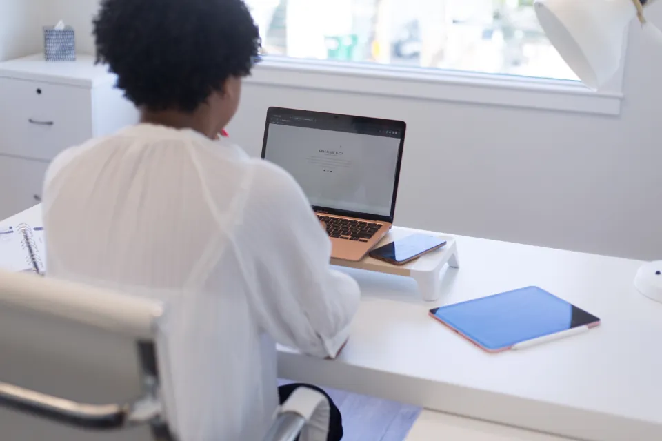 Personne vue de dos qui travaille sur un ordinateur portable à un bureau blanc, près d'une fenêtre.