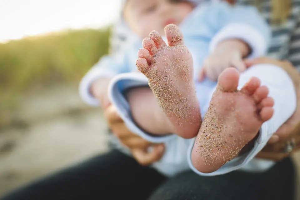 Gros plan sur les pieds d'un bébé couverts de sable, tenu dans les bras d'un adulte sur une plage.