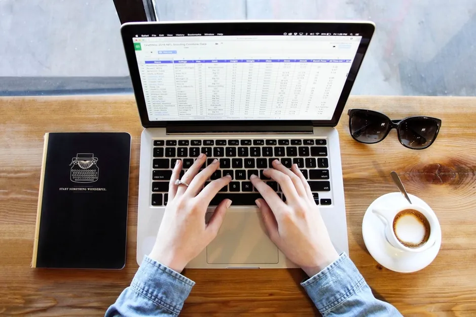 Mains sur le clavier d'un ordinateur portable affichant un tableur, avec carnet, lunettes de soleil et café sur un bureau en bois.
