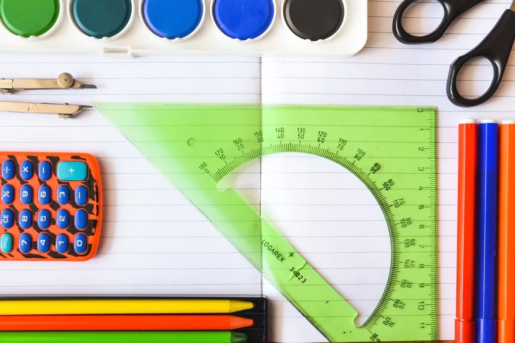 Fournitures scolaires vues de dessus : rapporteur, règle, crayons de couleur, ciseaux, calculatrice et godets de peinture.