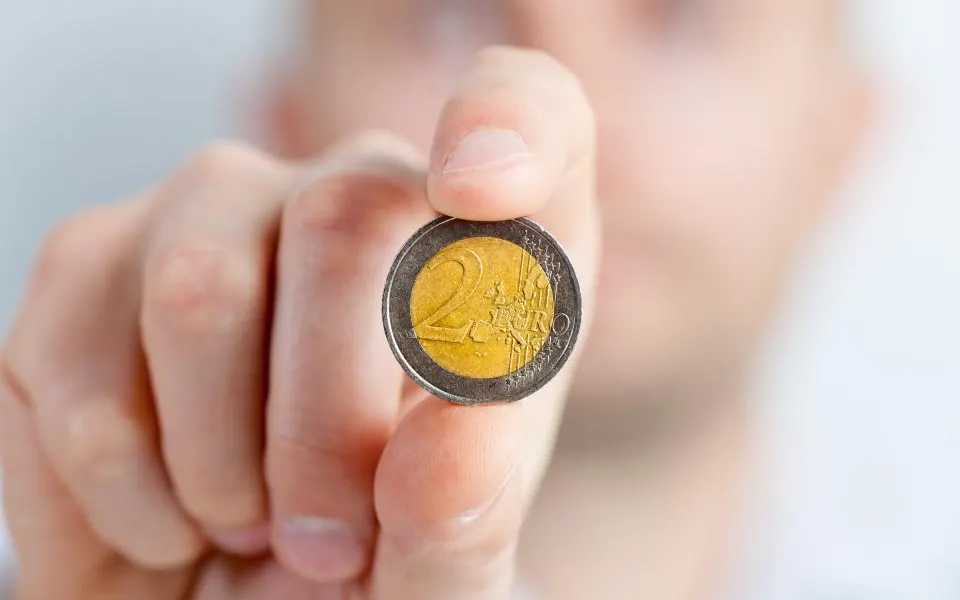 Main qui tient une pièce de 2 euros entre le pouce et l'index, au premier plan, sur un fond clair et flou.