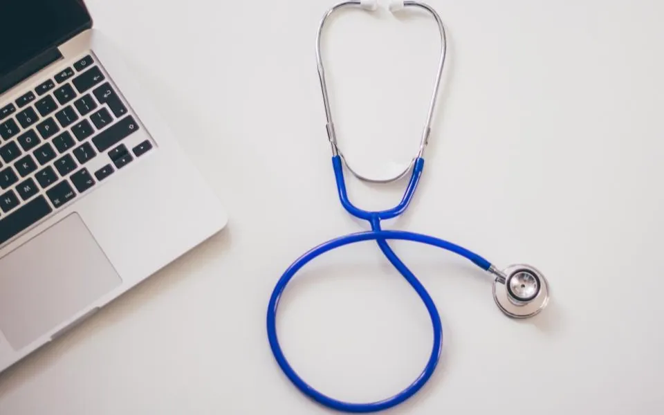 Stéthoscope bleu posé sur un bureau blanc, à côté d'un ordinateur portable.