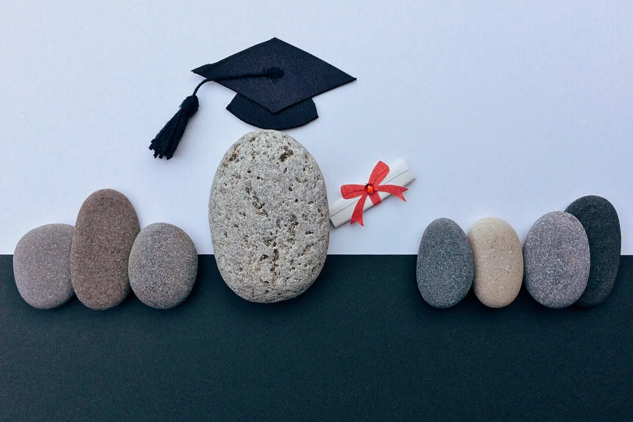 Toque de diplômé posée sur un galet, entourée de galets alignés et d'un diplôme roulé noué d'un ruban rouge.
