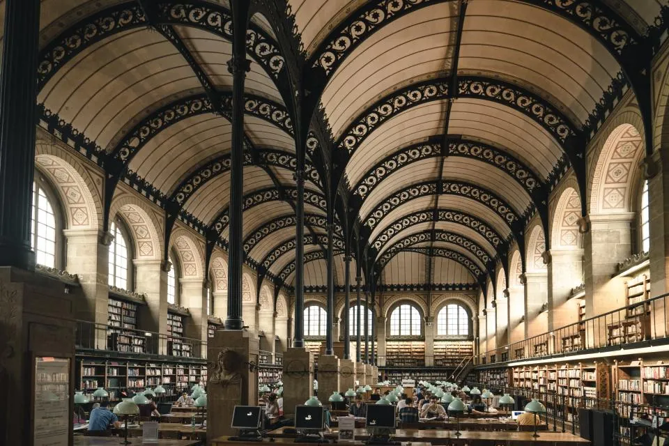 Grande salle de lecture d'une bibliothèque ancienne, sous une verrière à arches métalliques.