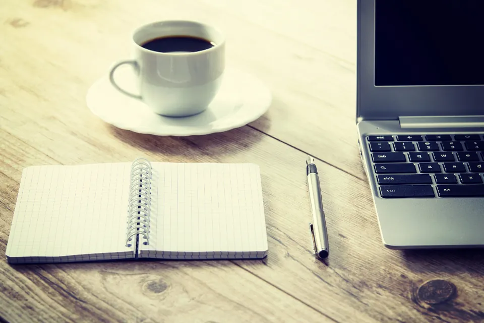 Tasse de café, carnet à spirale, stylo et ordinateur portable sur une table en bois.