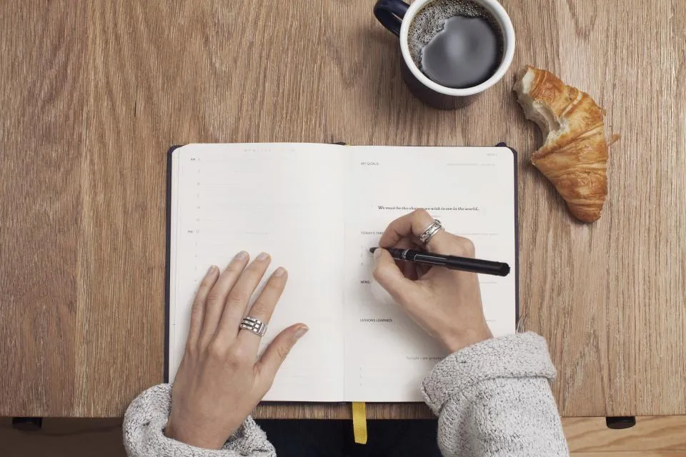 Main qui écrit dans un carnet ouvert sur une table en bois, tasse de café et croissant à côté.
