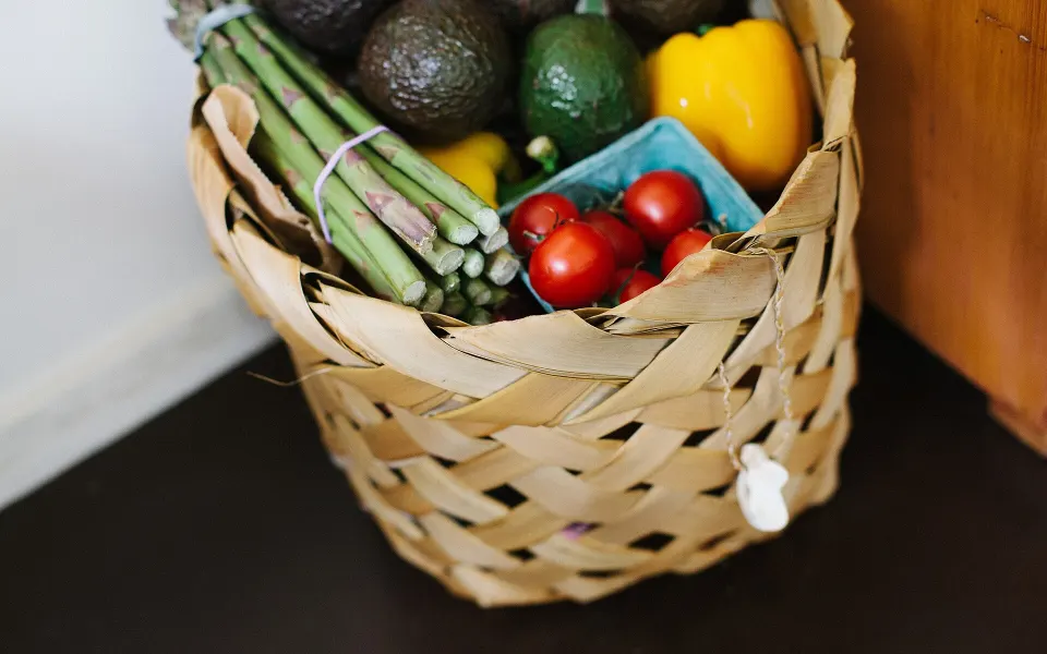 Panier en osier rempli d'asperges, d'avocats, de poivrons jaunes et d'une barquette de tomates cerises, posé au sol contre un meuble en bois.